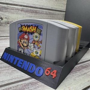 May include: A black 3D-printed Nintendo 64 game cartridge holder with the text "NINTENDO 64" in blue and red. The holder contains several grey and yellow game cartridges, including one labeled "Super Smash Bros."