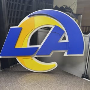 Pode incluir: Um logótipo tridimensional dos Los Angeles Rams com uma grande letra L e A azul integrada com uma forma de chifre amarelo curvo. O design é em camadas com um fundo branco, exibindo cores azul e amarelo arrojadas num formato estruturado.