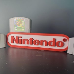 Peut inclure: Un panneau logo Nintendo rouge et blanc affiché horizontalement avec trois cartouches de jeu Nintendo 64 grises positionnées autour, incluant Mario Party 3, Super Mario 64 et Super Smash Bros, chacune avec des étiquettes de personnages colorées.