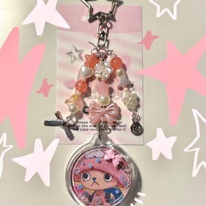 May include: A clear acrylic keychain featuring a cartoon character in a pink hat. The keychain has a star-shaped clasp, a beaded strand with pearls and flowers, and a pink bow. The text on the tag reads "Happy I...ke the unb..."