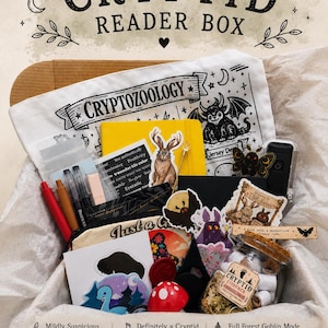 May include: A cardboard box filled with various items including a white tote bag labeled CRYPTOZOOLOGY, a yellow notebook, a glass jar labeled CRYPTID EVIDENCE, several stickers featuring creatures, pens, a red mushroom ornament, and a black pouch.