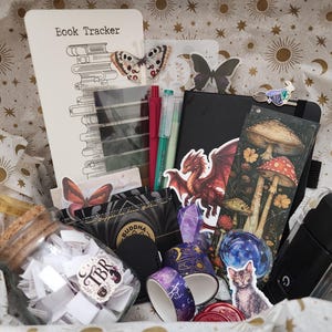Pode incluir: Uma cole&ccedil;&atilde;o de artigos de papelaria incluindo um cart&atilde;o Book Tracker, um caderno preto, um frasco de vidro cheio de pap&eacute;is com a etiqueta TBR, dois rolos de fita washi estampada, um adesivo de drag&atilde;o, um marcador de cogumelos e um adesivo de gato.