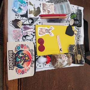 May include: A white tote bag featuring a black and white print, topped with a yellow notebook, a small canvas pouch with a floral Bigfoot graphic, several stickers, pens, a mushroom-shaped light, a glass jar, and various small stationery items.