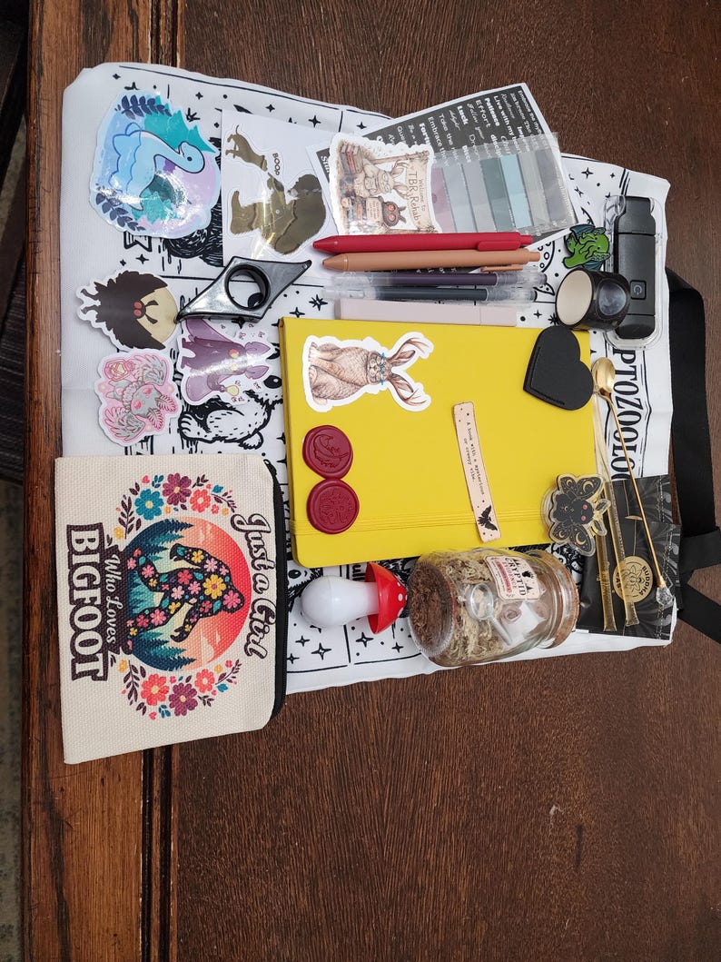 May include: A white tote bag featuring a black and white print, topped with a yellow notebook, a small canvas pouch with a floral Bigfoot graphic, several stickers, pens, a mushroom-shaped light, a glass jar, and various small stationery items.