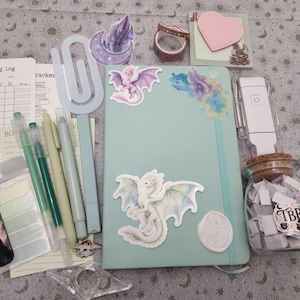 May include: A light green notebook decorated with dragon stickers, accompanied by a reading log, pens, a large paperclip, a glass jar filled with paper slips labeled TBR, a heart-shaped sticky note, a wax seal, and various stationery accessories.