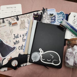 Puede incluir: Un estuche con cremallera con ilustraciones de gatos y el texto "Just a girl who loves cats" que contiene un cuaderno negro, dos rotuladores, un frasco de vidrio etiquetado "TBR" con papeles, pegatinas de gatos, una herramienta con forma de gato y registros de lectura.