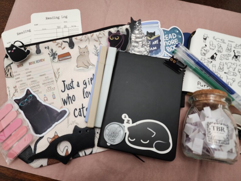 Puede incluir: Un estuche con cremallera con ilustraciones de gatos y el texto "Just a girl who loves cats" que contiene un cuaderno negro, dos rotuladores, un frasco de vidrio etiquetado "TBR" con papeles, pegatinas de gatos, una herramienta con forma de gato y registros de lectura.