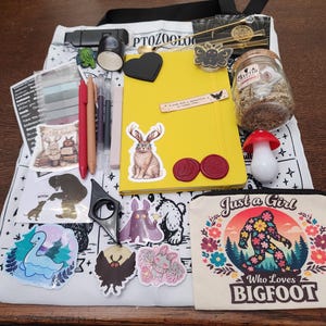 May include: A white tote bag featuring black illustrations and text, topped with a yellow notebook, a small glass jar with a cork lid, a mushroom-shaped item, a canvas pouch with the text Just a Girl Who Loves BIGFOOT, and various stickers.