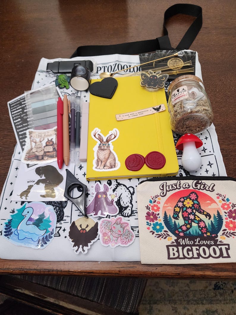 May include: A white tote bag featuring black illustrations and text, topped with a yellow notebook, a small glass jar with a cork lid, a mushroom-shaped item, a canvas pouch with the text Just a Girl Who Loves BIGFOOT, and various stickers.