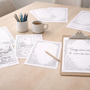 May include: A collection of coloring pages, pencils, and a clipboard with the text "Carry what helps. Leave the rest." are arranged on a light wood table. The coloring pages feature floral borders and whimsical illustrations, alongside a cup of tea.