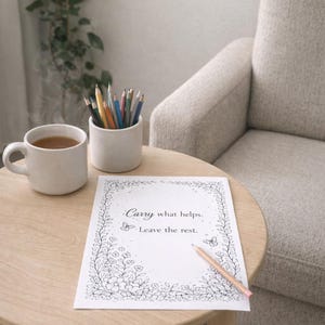May include: A serene scene featuring a round wooden table with a cup of tea, a small white ceramic container filled with colored pencils, and a coloring page with the text "Carry what helps. Leave the rest." A pencil rests on the page. A beige armchair is in the background.