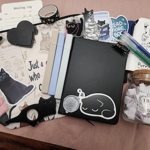 Puede incluir: Una colecci&oacute;n de art&iacute;culos de papeler&iacute;a que incluye un estuche con cremallera con tem&aacute;tica de gatos, un cuaderno negro con una pegatina de un gato durmiendo, tres marcadores pastel, un frasco de vidrio con papeles etiquetados TBR y clips.