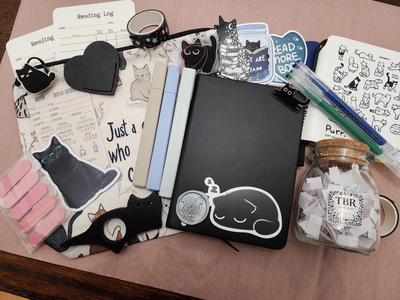 Puede incluir: Una colecci&oacute;n de art&iacute;culos de papeler&iacute;a que incluye un estuche con cremallera con tem&aacute;tica de gatos, un cuaderno negro con una pegatina de un gato durmiendo, tres marcadores pastel, un frasco de vidrio con papeles etiquetados TBR y clips.