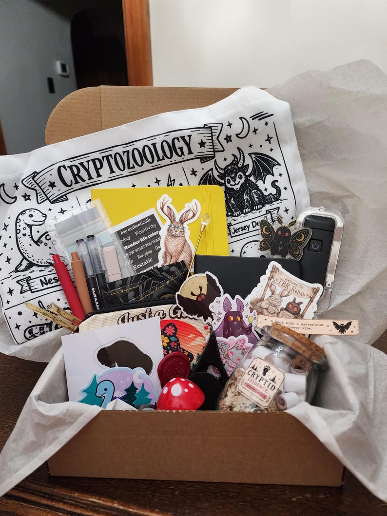 May include: A cardboard box filled with stationery and accessories, including a white pouch labeled CRYPTOZOOLOGY, a yellow notebook, pens, stickers featuring creatures, a glass jar labeled CRYPTID EVIDENCE, and a red mushroom-shaped object.