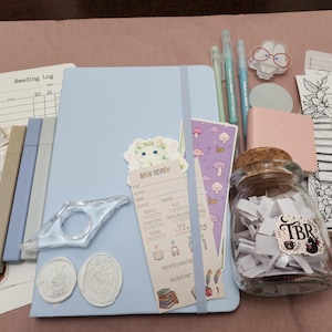 Puede incluir: Un cuaderno azul claro con banda el&aacute;stica, acompa&ntilde;ado de un frasco de vidrio lleno de tiras de papel blanco etiquetadas como TBR, una tarjeta de rese&ntilde;a de libros, un sujetap&aacute;ginas, tres bol&iacute;grafos claros, un marcap&aacute;ginas, pegatinas y dos sellos de cera.