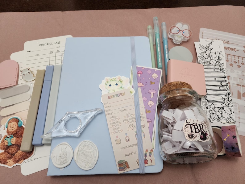 Puede incluir: Un cuaderno azul claro con banda el&aacute;stica, acompa&ntilde;ado de un frasco de vidrio lleno de tiras de papel blanco etiquetadas como TBR, una tarjeta de rese&ntilde;a de libros, un sujetap&aacute;ginas, tres bol&iacute;grafos claros, un marcap&aacute;ginas, pegatinas y dos sellos de cera.