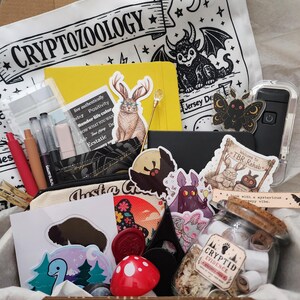 May include: A cardboard box filled with stationery and accessories, including a white tote bag with the text CRYPTOZOOLOGY, a yellow notebook, a glass jar labeled CRYPTID EVIDENCE, various stickers featuring creatures, a red mushroom object, and pens.