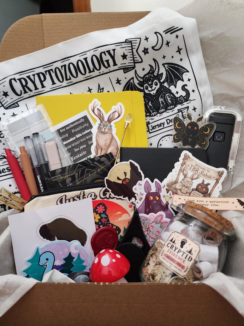 May include: A cardboard box filled with stationery and accessories, including a white tote bag with the text CRYPTOZOOLOGY, a yellow notebook, a glass jar labeled CRYPTID EVIDENCE, various stickers featuring creatures, a red mushroom object, and pens.