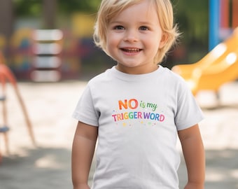 May include: White t-shirt with the text "NO is my TRIGGER WORD" in colorful letters. The shirt is worn by a smiling child with blonde hair. The background is a blurred outdoor playground.