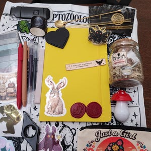 May include: A yellow notebook sits on a white tote bag printed with the word CRYPTOZOOLOGY. The collection includes a glass jar labeled CRYPTID EVIDENCE, a red mushroom figurine, pens, stickers of a jackalope and moth, wax seals, and a small gold spoon.