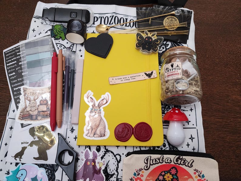May include: A yellow notebook sits on a white tote bag printed with the word CRYPTOZOOLOGY. The collection includes a glass jar labeled CRYPTID EVIDENCE, a red mushroom figurine, pens, stickers of a jackalope and moth, wax seals, and a small gold spoon.