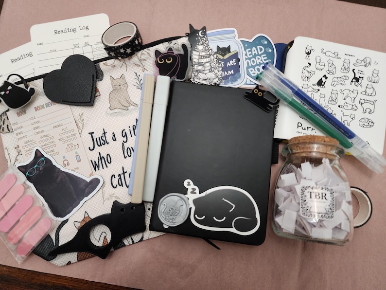 Puede incluir: Una colecci&oacute;n de art&iacute;culos de papeler&iacute;a que incluye un cuaderno negro, un estuche con motivos de gatos, varias pegatinas de gatos, un frasco de vidrio lleno de peque&ntilde;os papeles con la etiqueta TBR, dos marcadores, cinta washi y hojas de registro.