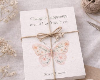 May include: A stack of Sobriety Affirmation Cards tied with twine, featuring a pink floral butterfly illustration. The top card reads, Change is happening, even if I can't see it yet. and Slow still counts. Additional cards and a sticker are visible.