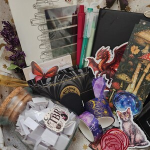 Pode incluir: Uma cole&ccedil;&atilde;o de artigos de papelaria e passatempos, incluindo um frasco de vidro cheio de pap&eacute;is, dois rolos de fita adesiva decorada, um caderno preto, um marcador de livros com ilustra&ccedil;&otilde;es de cogumelos, um autocolante de drag&atilde;o e um de gato.
