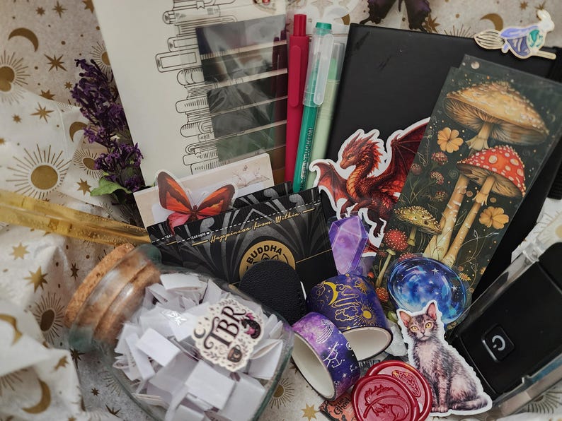 Pode incluir: Uma cole&ccedil;&atilde;o de artigos de papelaria e passatempos, incluindo um frasco de vidro cheio de pap&eacute;is, dois rolos de fita adesiva decorada, um caderno preto, um marcador de livros com ilustra&ccedil;&otilde;es de cogumelos, um autocolante de drag&atilde;o e um de gato.