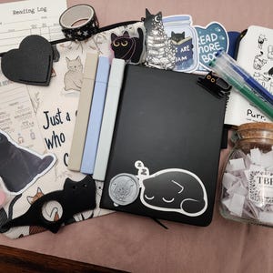 Puede incluir: Un conjunto de art&iacute;culos de papeler&iacute;a que incluye un cuaderno negro, tres marcadores pastel, un frasco de vidrio con tapa de corcho etiquetado TBR con papeles, un estuche con motivos de gatos, varias pegatinas de gatos y dos bol&iacute;grafos.