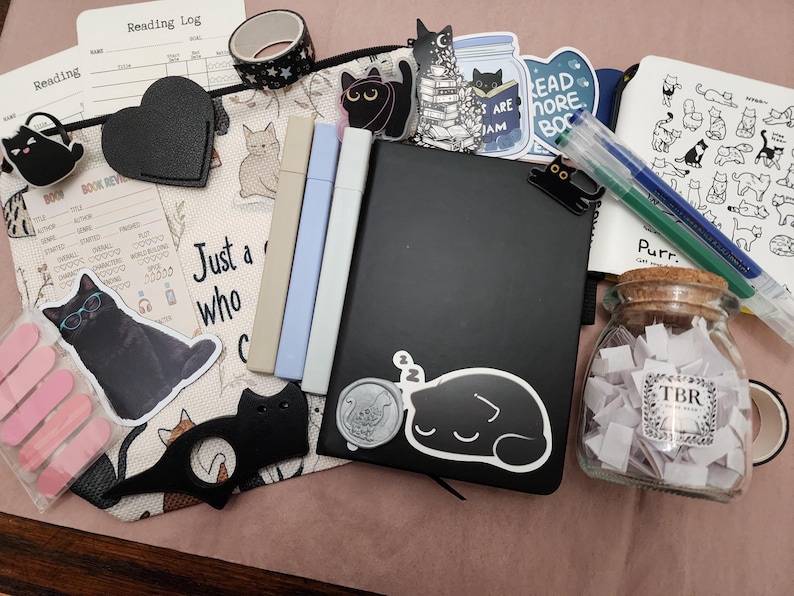 Puede incluir: Un conjunto de art&iacute;culos de papeler&iacute;a que incluye un cuaderno negro, tres marcadores pastel, un frasco de vidrio con tapa de corcho etiquetado TBR con papeles, un estuche con motivos de gatos, varias pegatinas de gatos y dos bol&iacute;grafos.