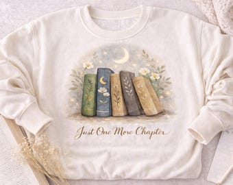 May include: Cream-colored sweatshirt with a watercolor illustration of a stack of books. The books are in shades of green, blue, and brown, with floral and celestial accents. The text "Just One More Chapter" is written below the books.