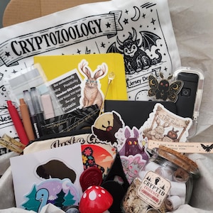 May include: A cardboard box filled with stationery and accessories, including a white tote bag with the text CRYPTOZOOLOGY, a yellow notebook, various stickers featuring creatures, a glass jar labeled CRYPTID EVIDENCE, and a red mushroom object.