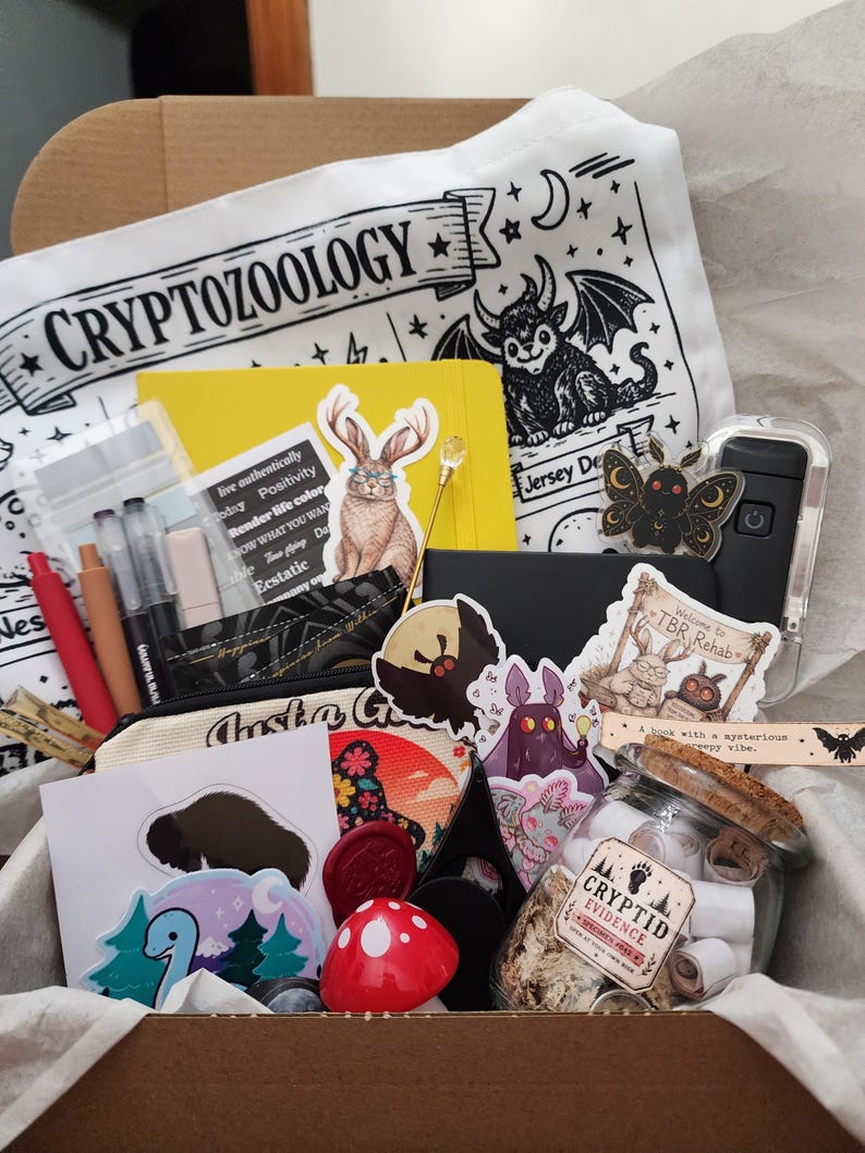 May include: A cardboard box filled with stationery and accessories, including a white tote bag with the text CRYPTOZOOLOGY, a yellow notebook, various stickers featuring creatures, a glass jar labeled CRYPTID EVIDENCE, and a red mushroom object.