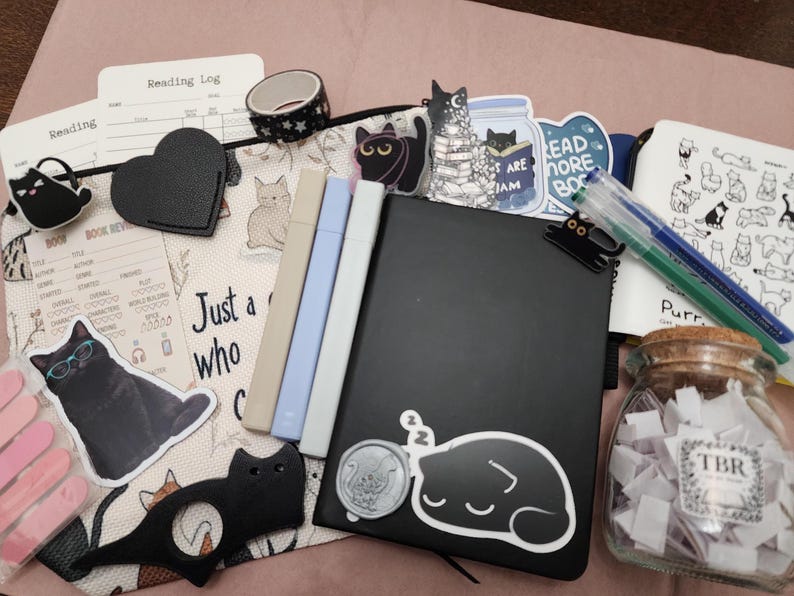Puede incluir: Un conjunto de art&iacute;culos de papeler&iacute;a que incluye un cuaderno negro con una pegatina de un gato durmiendo, tres marcadores pastel, un frasco de vidrio lleno de peque&ntilde;os papeles blancos etiquetados TBR, pegatinas de gatos, un clip negro en forma de coraz&oacute;n y un registro de lectura.