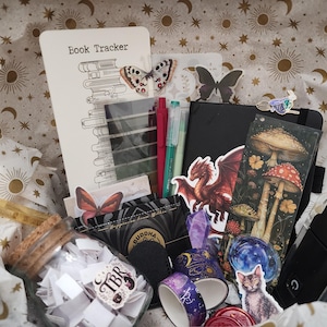 Pode incluir: Uma cole&ccedil;&atilde;o de artigos de papelaria incluindo um cart&atilde;o Book Tracker, um caderno preto, um marcador de p&aacute;gina com ilustra&ccedil;&otilde;es de cogumelos, um pote de vidro com pap&eacute;is, dois rolos de fita washi decorada, canetas e adesivos de drag&atilde;o e gato.