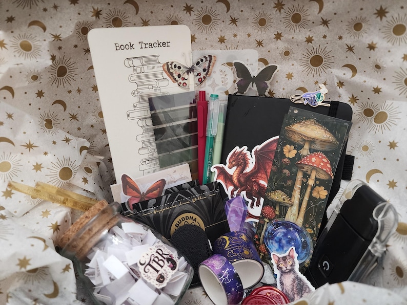 Pode incluir: Uma cole&ccedil;&atilde;o de artigos de papelaria incluindo um cart&atilde;o Book Tracker, um caderno preto, um marcador de p&aacute;gina com ilustra&ccedil;&otilde;es de cogumelos, um pote de vidro com pap&eacute;is, dois rolos de fita washi decorada, canetas e adesivos de drag&atilde;o e gato.