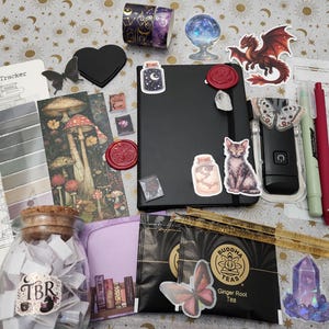 Pode incluir: Um caderno preto decorado com v&aacute;rios autocolantes, incluindo um gato, um frasco e um drag&atilde;o, acompanhado por um frasco de vidro com a etiqueta TBR, dois sacos de ch&aacute;, um marcador com ilustra&ccedil;&otilde;es de cogumelos, uma folha de registo e canetas.