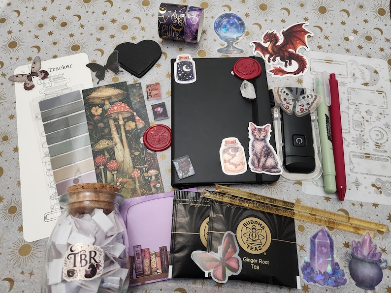 Pode incluir: Um caderno preto decorado com v&aacute;rios autocolantes, incluindo um gato, um frasco e um drag&atilde;o, acompanhado por um frasco de vidro com a etiqueta TBR, dois sacos de ch&aacute;, um marcador com ilustra&ccedil;&otilde;es de cogumelos, uma folha de registo e canetas.