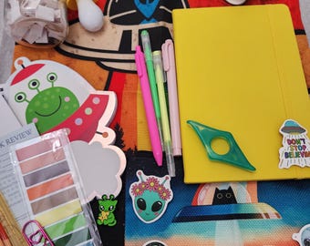 May include: A collection of stationery items including a yellow notebook, four pens, a glass jar with paper slips, two rolls of washi tape, a green ring-shaped tool, and various alien-themed stickers on a red and black tote bag background.
