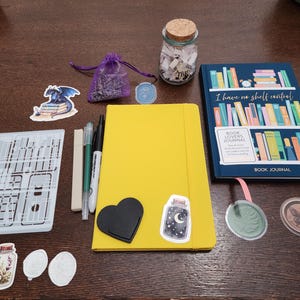 Puede incluir: Un conjunto de art&iacute;culos de papeler&iacute;a que incluye un cuaderno amarillo con pegatinas, un diario azul para lectores, un frasco de vidrio con tapa de corcho, una bolsa morada, tres bol&iacute;grafos, una plantilla y varias pegatinas decorativas.