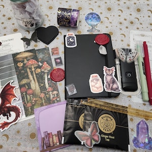Pode incluir: Um caderno preto decorado com autocolantes de um gato, um frasco e um selo de cera, rodeado por um frasco de vidro, fita adesiva, um marcador de cogumelos, um autocolante de drag&atilde;o vermelho, duas saquetas de ch&aacute;, uma r&eacute;gua, canetas e papelaria.