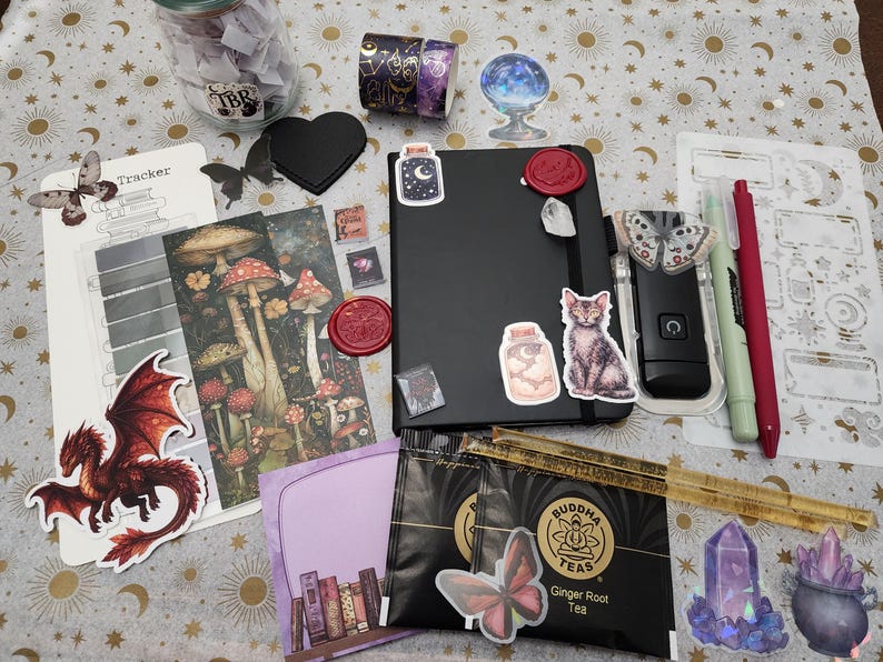 Pode incluir: Um caderno preto decorado com autocolantes de um gato, um frasco e um selo de cera, rodeado por um frasco de vidro, fita adesiva, um marcador de cogumelos, um autocolante de drag&atilde;o vermelho, duas saquetas de ch&aacute;, uma r&eacute;gua, canetas e papelaria.