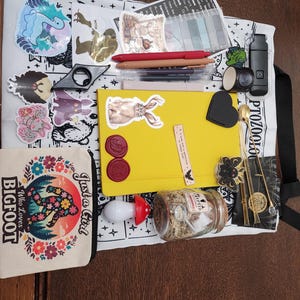 May include: A white tote bag featuring black illustrations and text, topped with a yellow notebook, a small canvas pouch with a Bigfoot graphic, pens, stickers, a glass jar with moss, a mushroom-shaped light, and various stationery accessories.