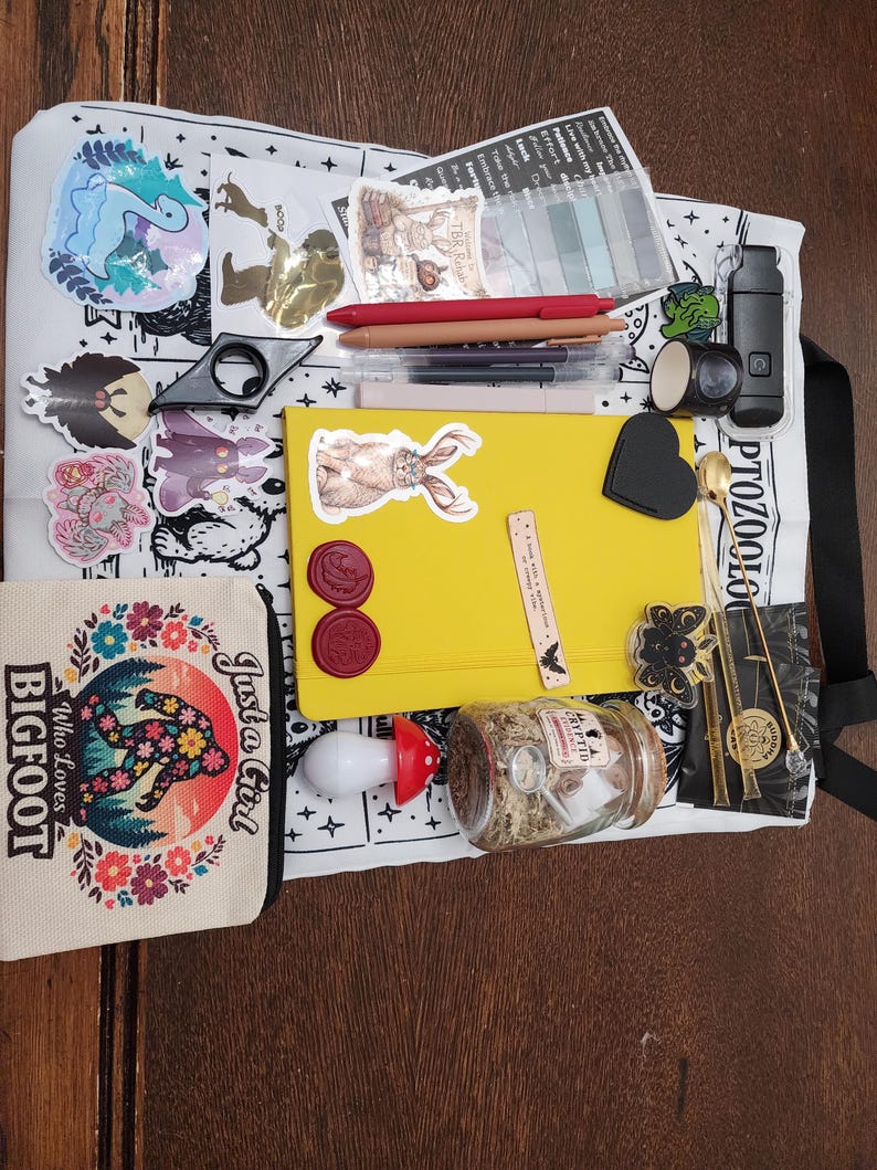 May include: A white tote bag featuring black illustrations and text, topped with a yellow notebook, a small canvas pouch with a Bigfoot graphic, pens, stickers, a glass jar with moss, a mushroom-shaped light, and various stationery accessories.