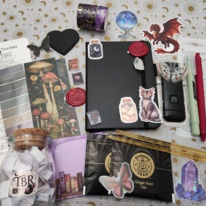 Pode incluir: Um conjunto de artigos de papelaria incluindo um caderno preto com autocolantes, um frasco de vidro com a etiqueta TBR cheio de tiras de papel, duas saquetas de ch&aacute; Buddha Teas Ginger Root, um marcador com ilustra&ccedil;&otilde;es de cogumelos, uma r&eacute;gua, canetas e v&aacute;rios autocolantes.