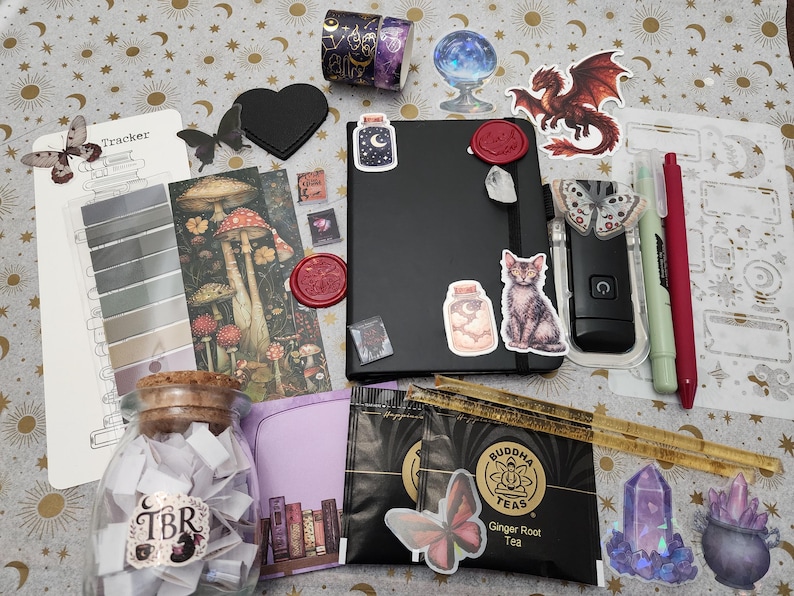 Pode incluir: Um conjunto de artigos de papelaria incluindo um caderno preto com autocolantes, um frasco de vidro com a etiqueta TBR cheio de tiras de papel, duas saquetas de ch&aacute; Buddha Teas Ginger Root, um marcador com ilustra&ccedil;&otilde;es de cogumelos, uma r&eacute;gua, canetas e v&aacute;rios autocolantes.
