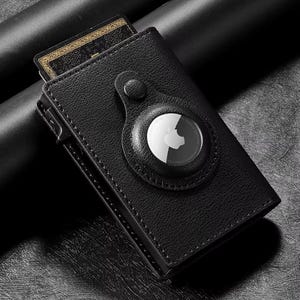 May include: Black leather wallet with a card peeking out. The wallet has a circular AirTag holder attached. The AirTag is white with a black Apple logo. The wallet is closed and has a button closure.