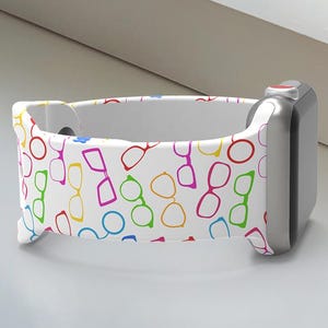 May include: White Apple Watch band with a colorful glasses pattern. The band is paired with a silver Apple Watch. A white Apple Watch box with the word "WATCH" is in the background.