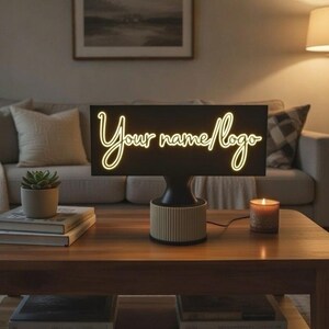 Könnte beinhalten: Eine schwarz-beige Tischlampe mit dem Text "Your name/logo" in Schreibschrift. Die Lampe steht auf einem Couchtisch aus Holz mit Büchern, einer Sukkulente und einer brennenden Kerze. Im Hintergrund sind ein Sofa und eine Lampe zu sehen.